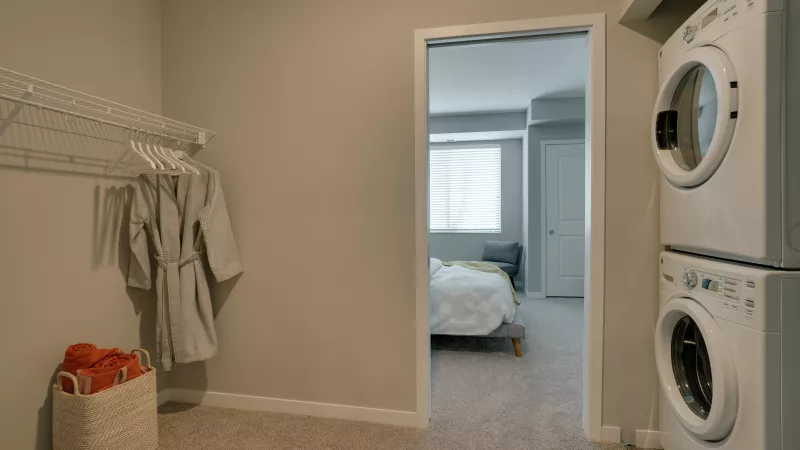 Stacked washer/dryer in a neutral laundry closet. Wire shelf, robe, and red towels in a basket. Bedroom visible through doorway.