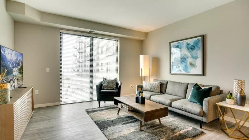 Modern living room with a grey sofa, wooden coffee table, armchair, TV, and abstract art, facing large sliding glass doors.
