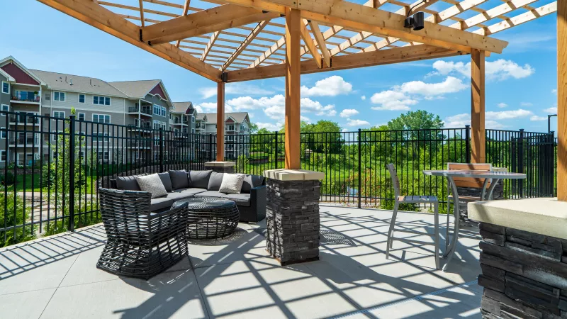 Outdoor patio with a wooden trellis, comfortable seating, and a table, overlooking a green landscape and apartment buildings.