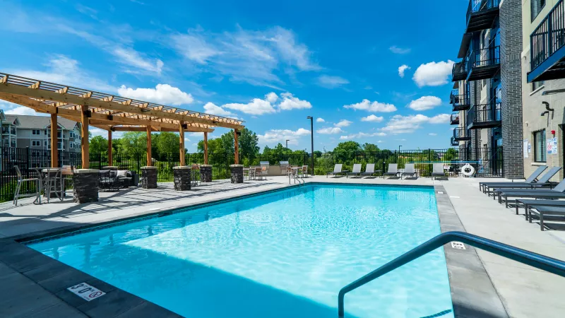 Outdoor swimming pool with lounge chairs, a wooden pergola seating area, and a modern apartment building under a blue sky.
