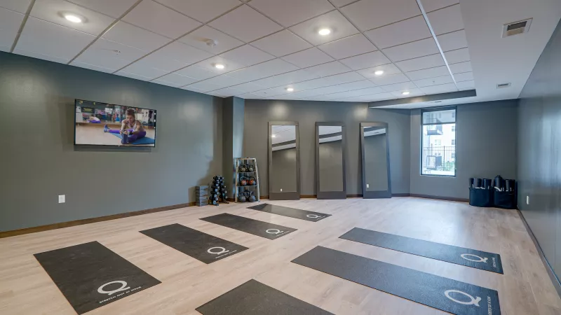 Bright yoga studio with grey walls, light wood floor, multiple mats, tall mirrors, and a TV displaying a workout.