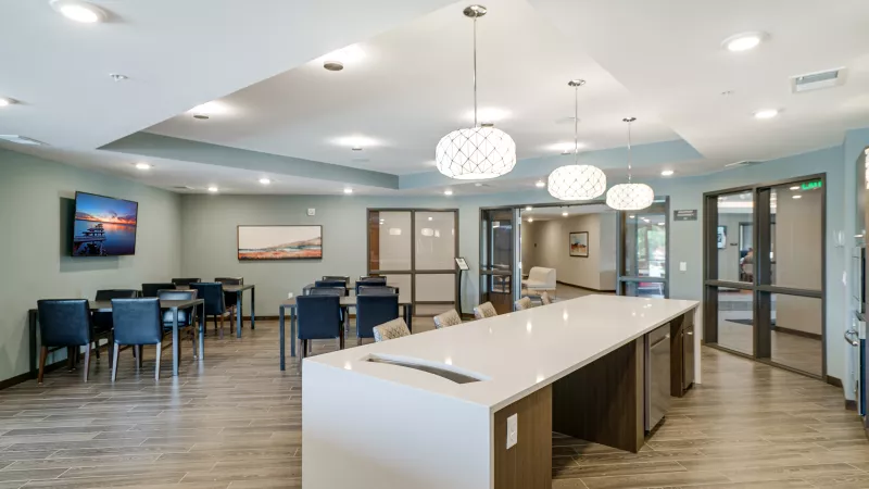 Modern common room with a long white kitchen island, dining tables with chairs, and a wall-mounted TV.