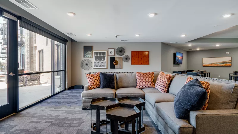 Modern lounge with grey sectional sofa, orange and blue pillows, hexagonal tables, wall art, and large windows.