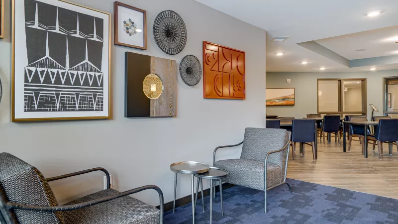 Modern lounge with two armchairs, a side table, and diverse wall art. Blue patterned carpet and a dining area in the background.