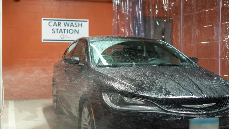 Black car covered in water and suds inside a car wash bay with an orange brick wall and "CAR WASH STATION" sign.