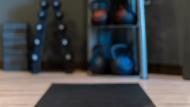 Photograph of a black yoga mat on a light floor in a home gym, with blurred kettlebells and dumbbells in the background.