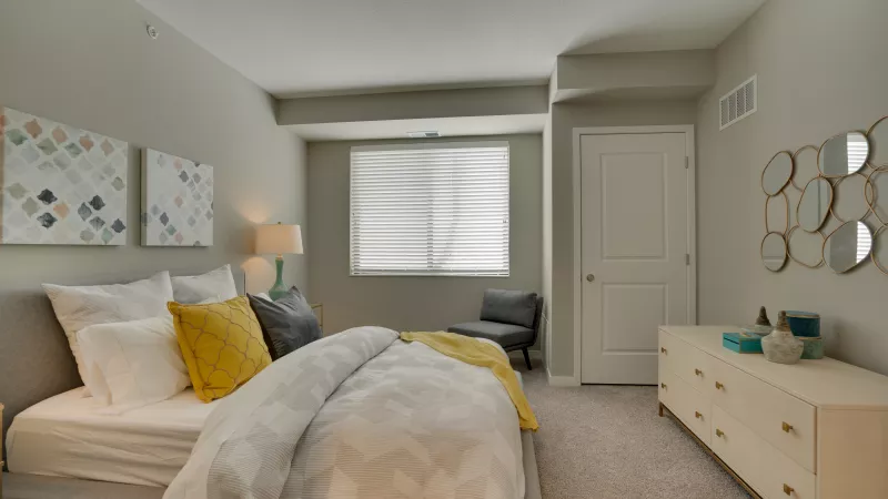 Modern bedroom with gray walls, bed with white and yellow bedding, dresser, and wall decor.