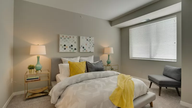 Modern bedroom with gray bed, yellow throw, turquoise lamps, abstract art, and gray armchair by a window.
