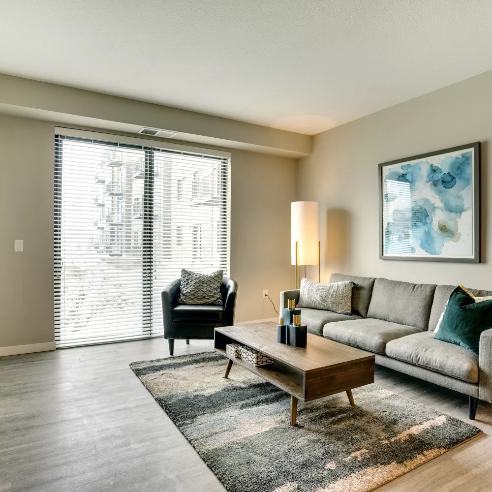 Modern living room with a grey sofa, wooden coffee table, armchair, TV, and abstract art, facing large sliding glass doors.