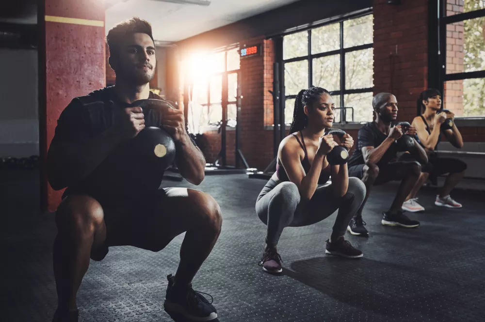 Four individuals focused, performing kettlebell squats together in a sunlit gym.