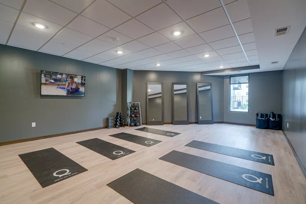 Bright yoga studio with grey walls, light wood floor, multiple mats, tall mirrors, and a TV displaying a workout.